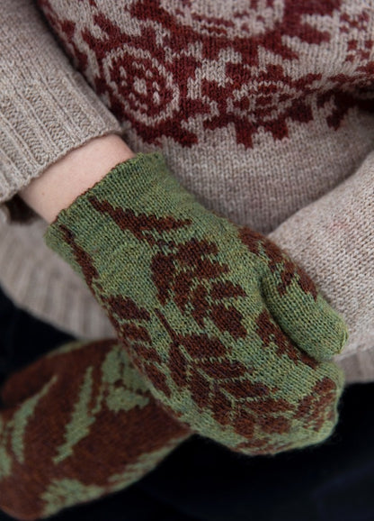 Beware the Frogs Mittens: green and brown leaf-patterned knit mittens shown with a beige sweater featuring red and white chest design.