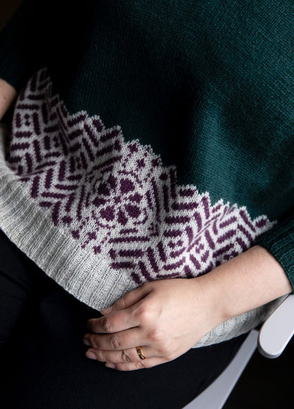 Compass Rose Sweater: dark green knit with purple and white compass rose motifs near hem, shown in a seated close-up.