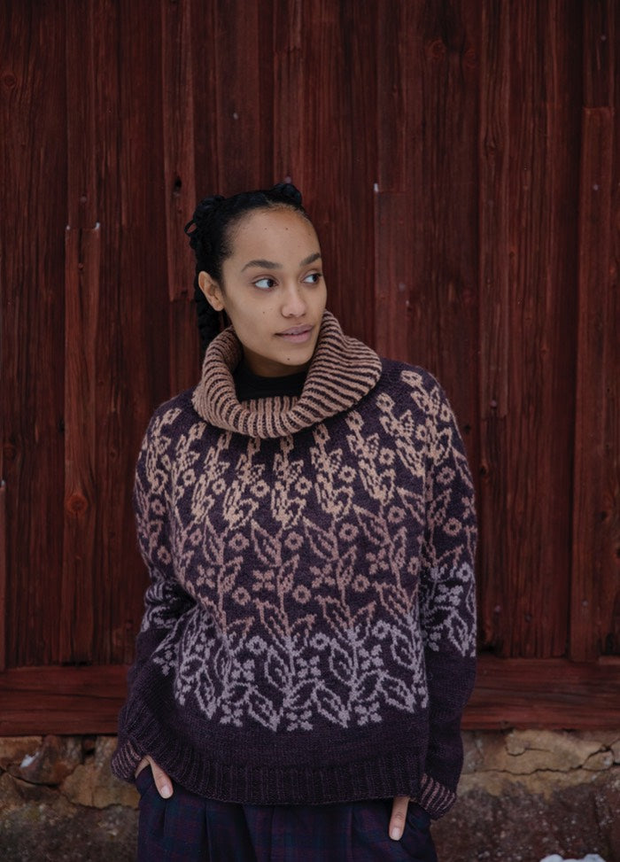 Deep Roots Sweater: dark knit with light root-like motifs and a high ribbed collar, shown against a reddish wooden backdrop.