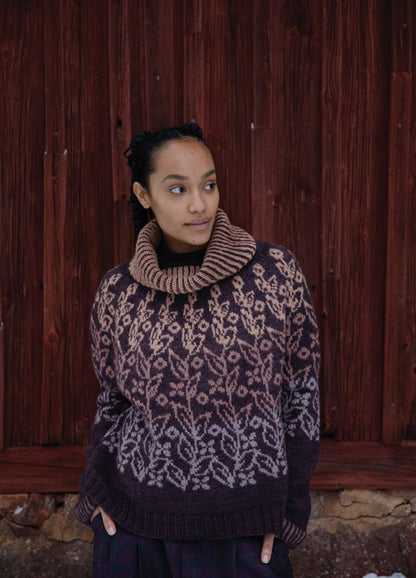 Deep Roots Sweater: dark knit with light root-like motifs and a high ribbed collar, shown against a reddish wooden backdrop.