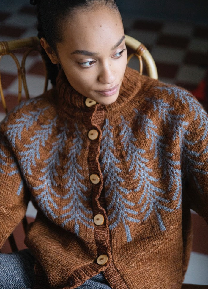 Fox Den Cardigan: brown knit with blue branch motifs and wooden buttons, shown seated on a woven-back chair.