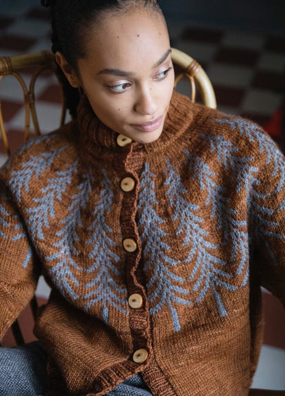Fox Den Cardigan: brown knit with blue branch motifs and wooden buttons, shown seated on a woven-back chair.