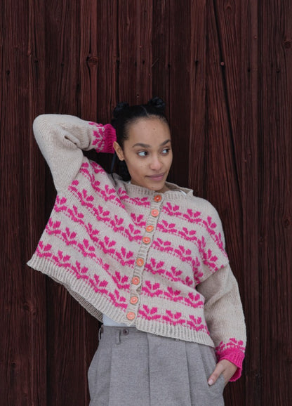 Garden Lupin Cardigan: light knit with pink lupin flower motifs in rows, featuring pink cuffs and light brown buttons, set against wooden planks.