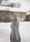 Like a Ram Cardigan: long thick knit with zigzag pattern, worn outdoors in a snowy setting near a house and trees.