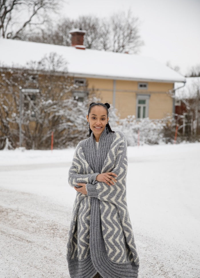 Like a Ram Cardigan: long thick knit with zigzag pattern, worn outdoors in a snowy setting near a house and trees.