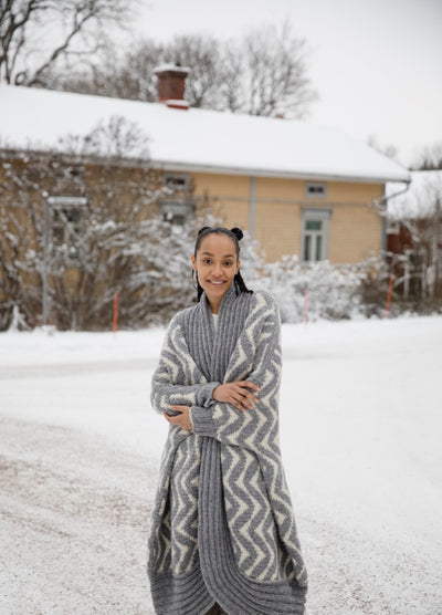 Like a Ram Cardigan: long thick knit with zigzag pattern, worn outdoors in a snowy setting near a house and trees.