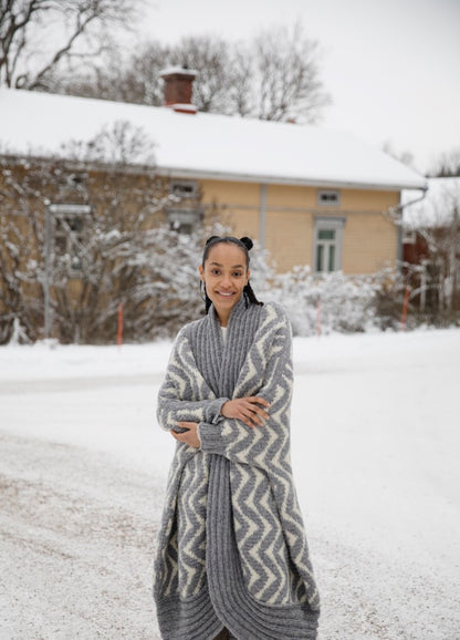 Like a Ram Cardigan: long thick knit with zigzag pattern, worn outdoors in a snowy setting near a house and trees.