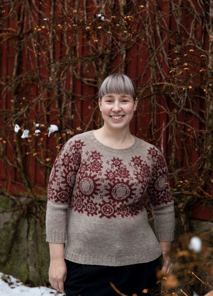 Manual Labour Sweater: beige knit with red geometric and circular motifs across the chest, set against a red wooden wall with bare branches.