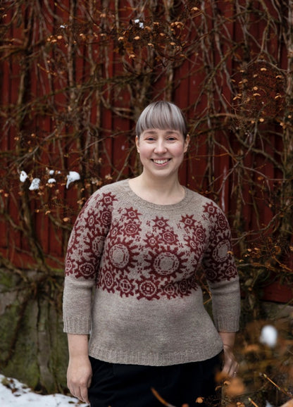 Manual Labour Sweater: beige knit with red geometric and circular motifs across the chest, set against a red wooden wall with bare branches.