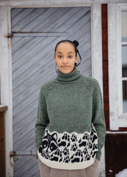 Rascal Spirit Sweater: green high-neck knit with black and white abstract animal print near hem, shown against a wooden door.
