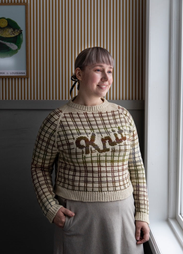 Vintage Flannel Sweater: checkered knit in brown, beige, and green with 'Knit' across chest, set against striped wallpaper and framed art.