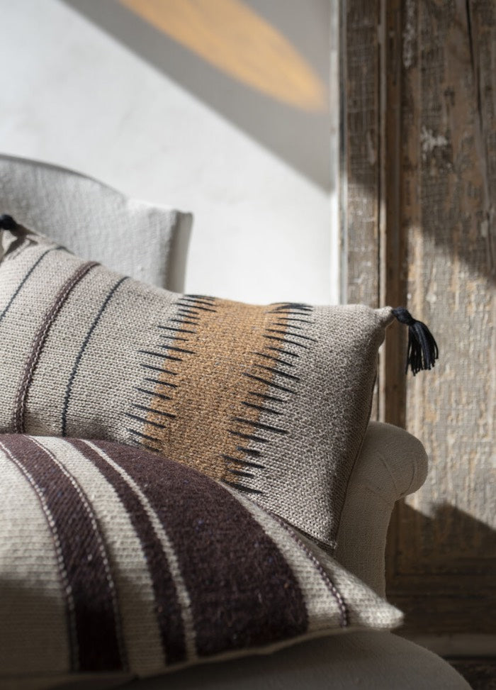 Cozy armchair with two woolen pillows; one with brown zigzag and black tassels, the other striped. Warm sunlight and rustic wooden backdrop.