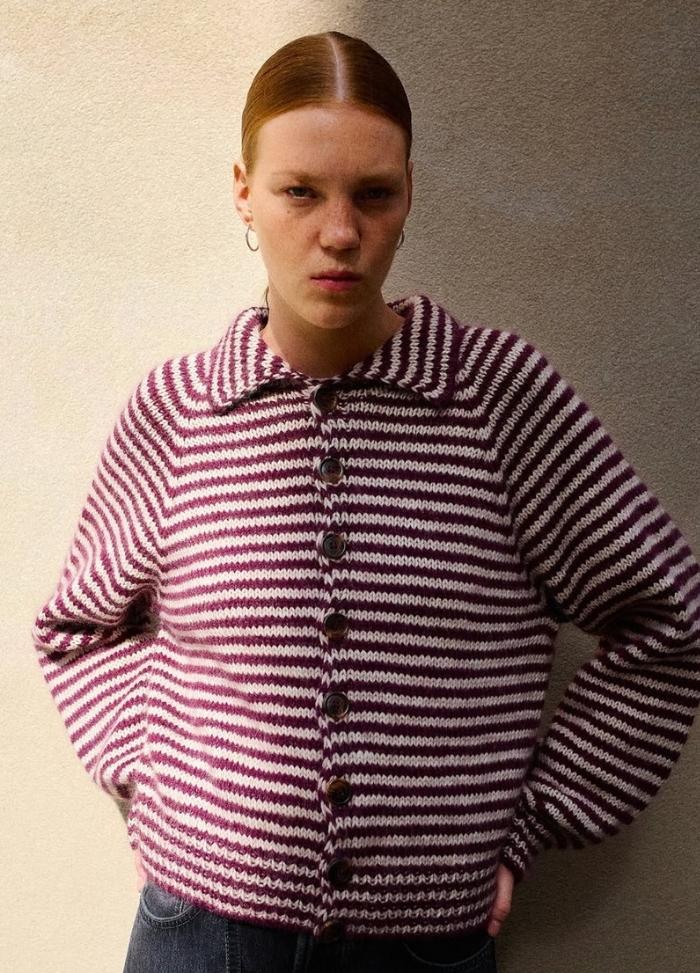 A person wearing a red and white striped sweater stands against a neutral wall, giving a serious expression. The sweater has large buttons and a wide collar.