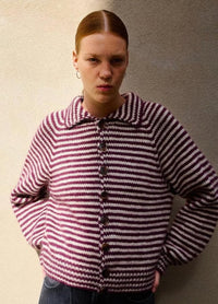 A person wearing a red and white striped sweater stands against a neutral wall, giving a serious expression. The sweater has large buttons and a wide collar.