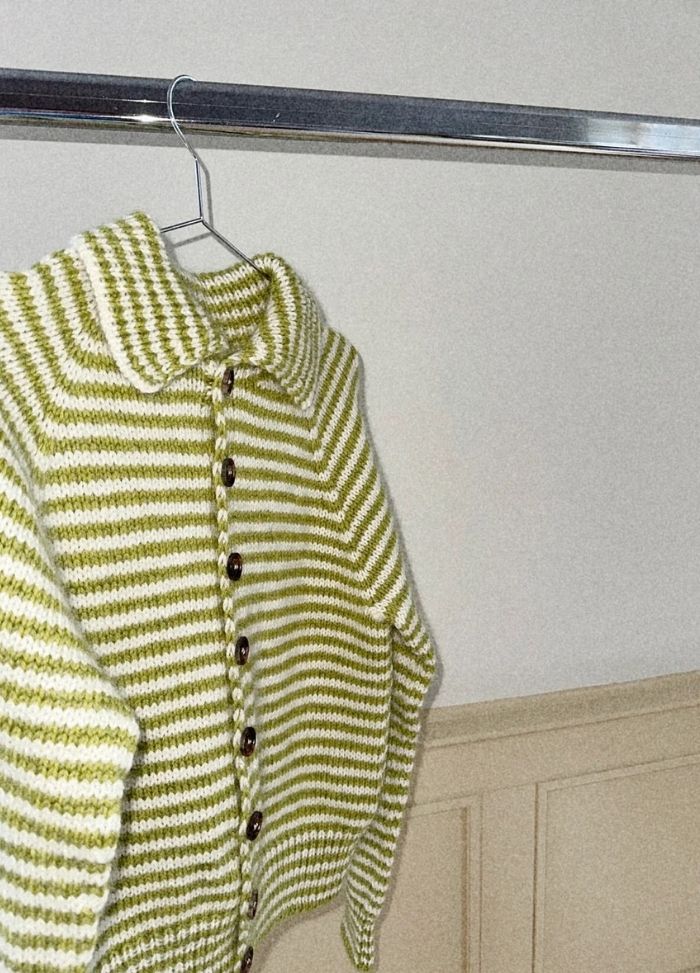 A cozy white and green striped knit cardigan hangs on a metal rack, featuring a collared design with dark buttons, exuding a warm, inviting feel.