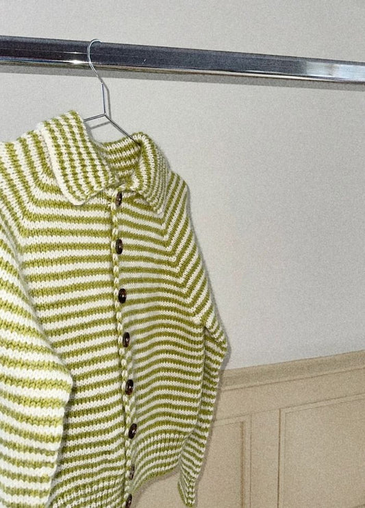 A cozy white and green striped knit cardigan hangs on a metal rack, featuring a collared design with dark buttons, exuding a warm, inviting feel.