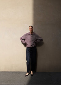 A woman stands against a beige wall, half in shadow. She wears a red and white striped sweater and dark pants. Her hands rest on her hips, conveying confidence.