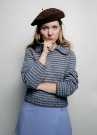 A person in a brown beret and striped sweater with blue and brown tones poses thoughtfully with a hand on their chin against a plain background.