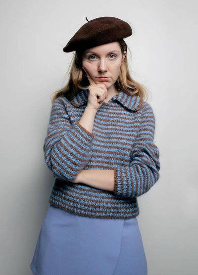 A person in a brown beret and striped sweater with blue and brown tones poses thoughtfully with a hand on their chin against a plain background.