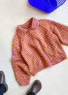Striped pink and orange knitted sweater with a wide collar on a light surface. Nearby, brown shoes and a blue basket are visible, conveying a cozy feel.