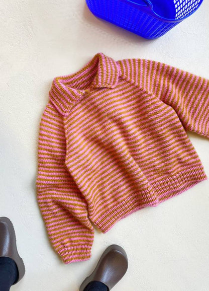 Striped pink and orange knitted sweater with a wide collar on a light surface. Nearby, brown shoes and a blue basket are visible, conveying a cozy feel.