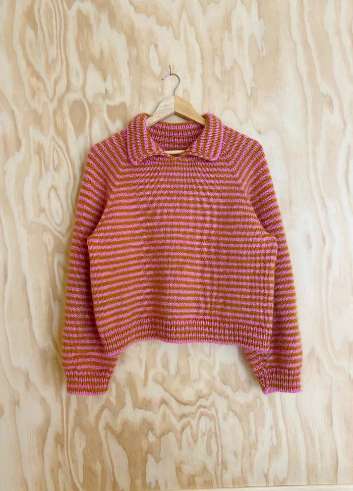 Striped orange and pink knit sweater with a collar, displayed on a wooden hanger against a light wood-panelled background, exuding a cozy vibe.