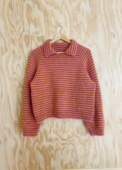 Striped orange and pink knit sweater with a collar, displayed on a wooden hanger against a light wood-panelled background, exuding a cozy vibe.