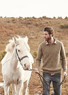 Person wearing the Suffolk Sweater, walking beside a white horse in a hilly outdoor landscape.