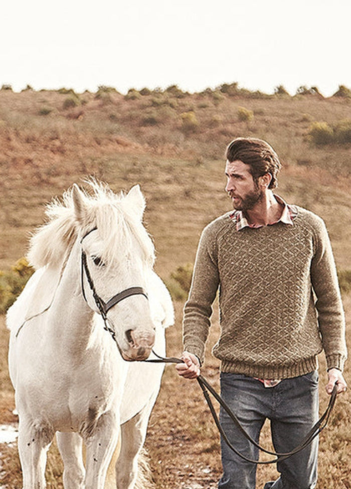 Person wearing the Suffolk Sweater, walking beside a white horse in a hilly outdoor landscape.