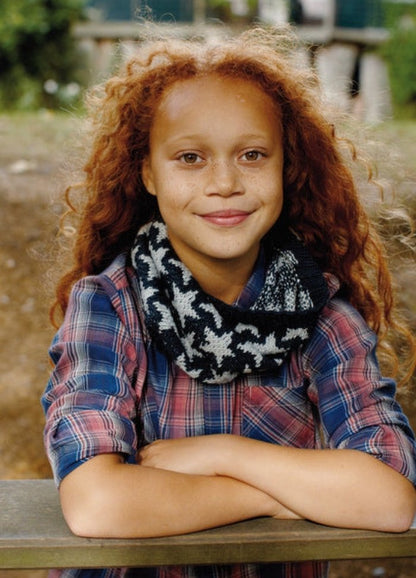 Child in Sundance Cowl with star motif over plaid shirt, outdoors with greenery.