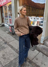 Woman in a cozy brown sweater and jeans stands on a city sidewalk, holding a dark coat.