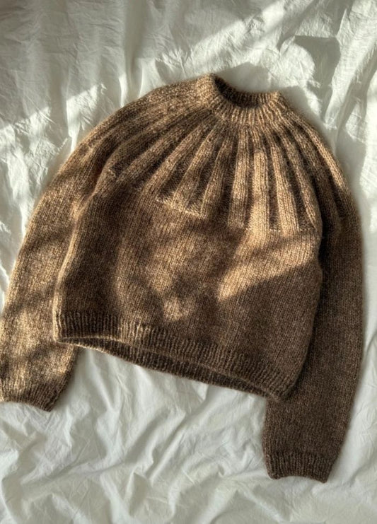 A cozy brown knit sweater with a ribbed neck and textured pattern is laid out on a softly crumpled white sheet, exuding a warm, autumnal feel.