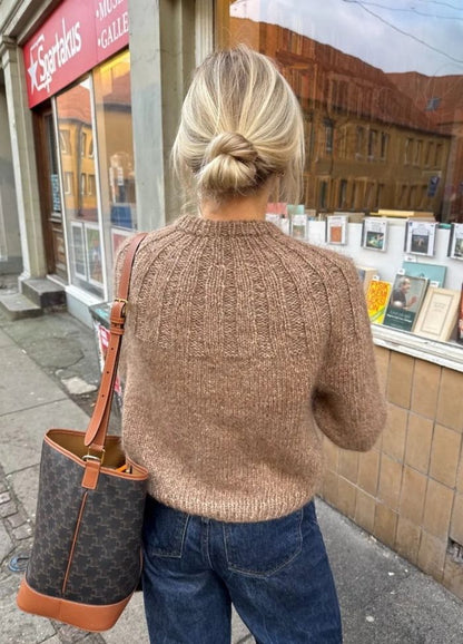 A woman with a blonde bun stands outside a bookstore, wearing a brown sweater and jeans.