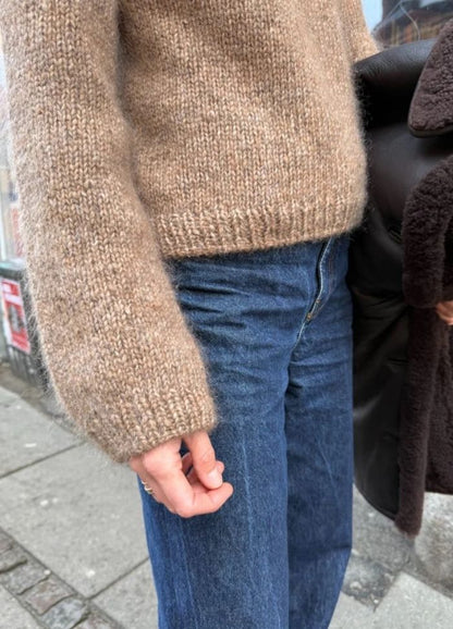 Person wearing a cozy beige knit sweater and blue jeans stands on a sidewalk. A dark brown coat is draped over one arm, suggesting cool weather.