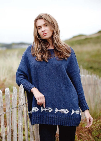 Young woman with long hair wearing a blue sweater with fishbone design stands by a wooden fence. Background features grassy dunes, creating a relaxed mood.