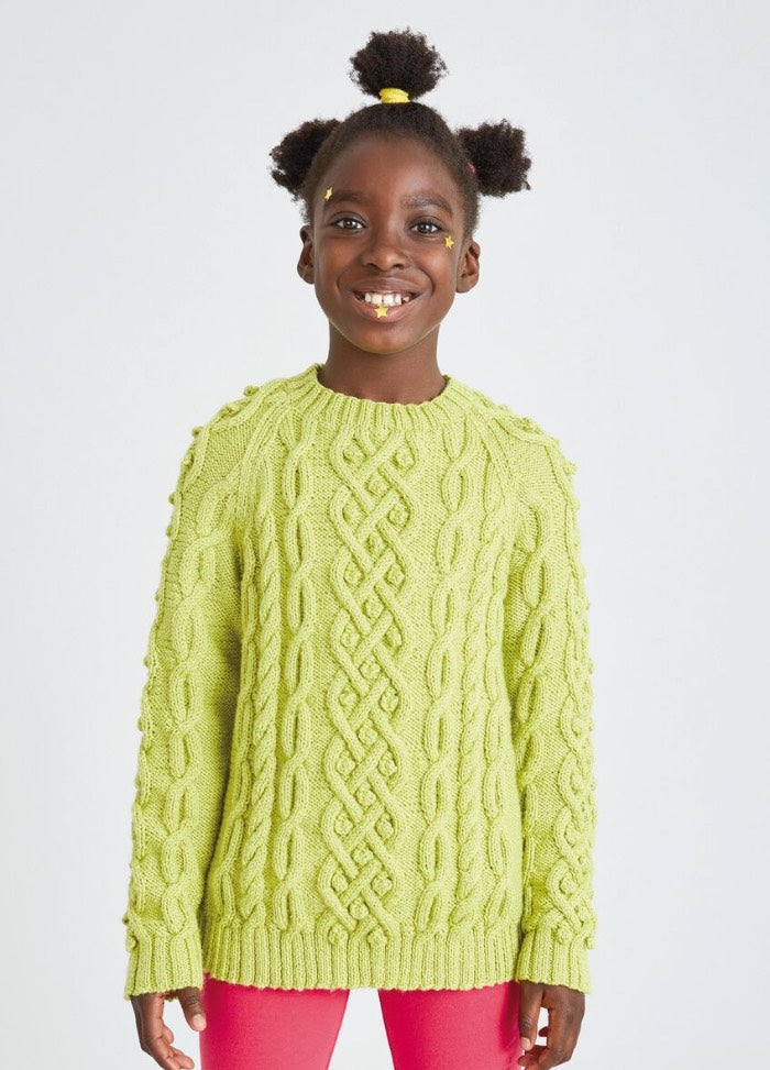 A smiling child wearing a bright yellow cable-knit sweater and red pants stands against a plain background. They convey a joyful and cheerful vibe.