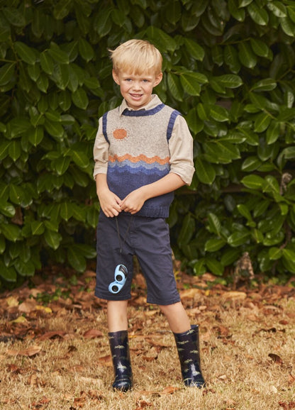 A smiling young boy stands in front of lush green foliage. He's wearing a colorful knit vest, navy shorts, and patterned rain boots, exuding a playful vibe.
