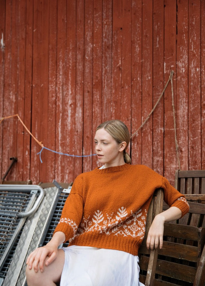 Suolaulu Sweater: orange knit with white snowflake motifs on the hem and sleeves, worn seated against a rustic red wooden wall.