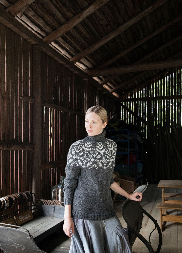 Suolaulu Talvi Sweater: dark grey knit with a detailed white yoke pattern, worn indoors in a rustic wooden setting.