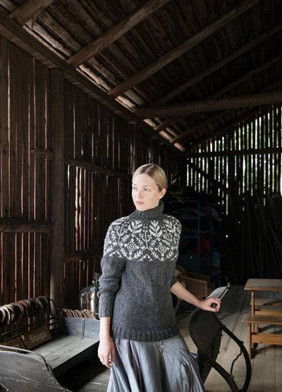 Suolaulu Talvi Sweater: dark grey knit with a detailed white yoke pattern, worn indoors in a rustic wooden setting.