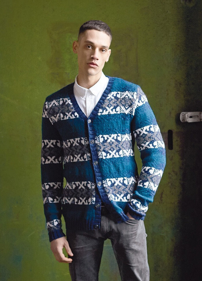 A person leans against a green wall, wearing a blue and white patterned cardigan with a white shirt underneath. They have a calm expression.