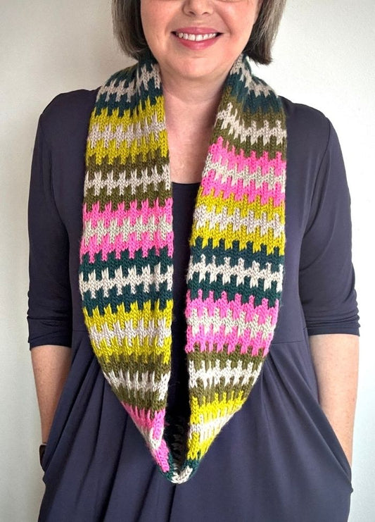 Smiling woman wearing a multicolored knitted scarf with stripes of green, pink, gray, and white, over a dark blue top. The mood is warm and cheerful.