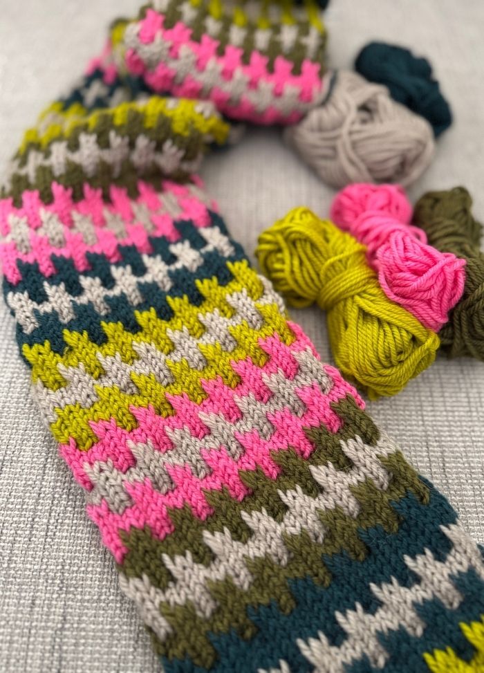 A colorful knitted scarf with a houndstooth pattern in pink, green, blue, and beige lies on a gray surface. Nearby are yarn balls in similar colors.