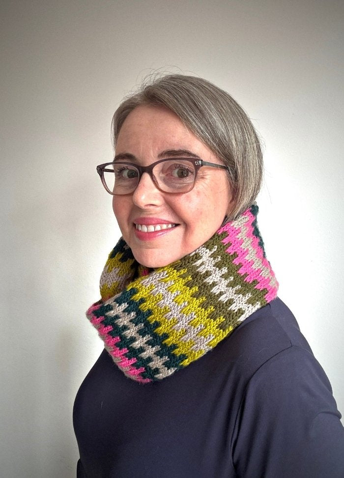 A smiling person with glasses and short gray hair wears a colorful knit scarf with pink, green, and yellow stripes over a dark top. Plain background.