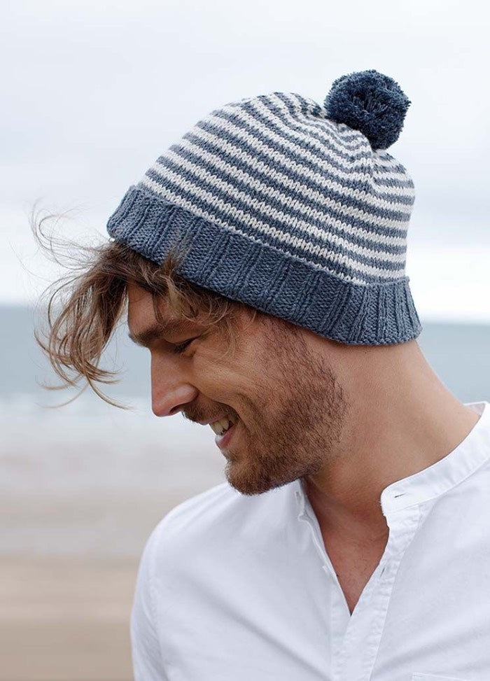 Man smiling and looking down, wearing a striped knit beanie with a pom-pom. He has tousled hair and a white shirt against a blurred beach background.