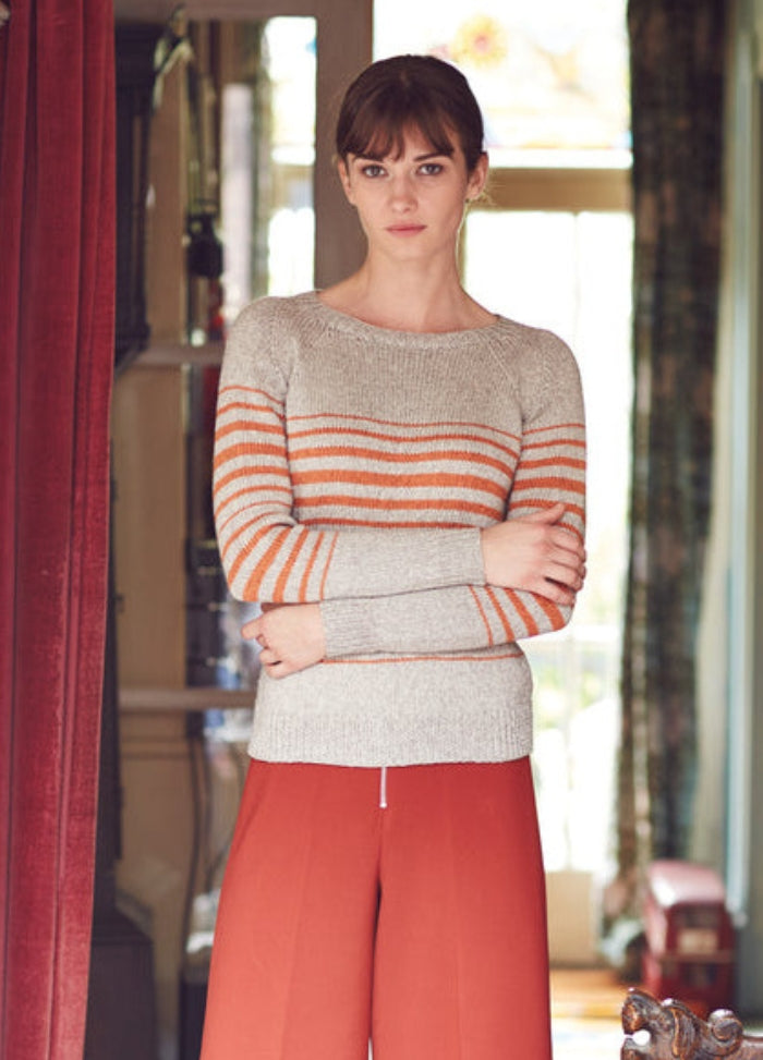 Model wearing the Tinley Sweater from the Rowan Softyak DK Collection by Marie Wallin, a light gray knit with horizontal orange and gray stripes across the chest and sleeves, paired with high-waisted red wide-leg pants, photographed indoors with a red curtain and natural light from a window.