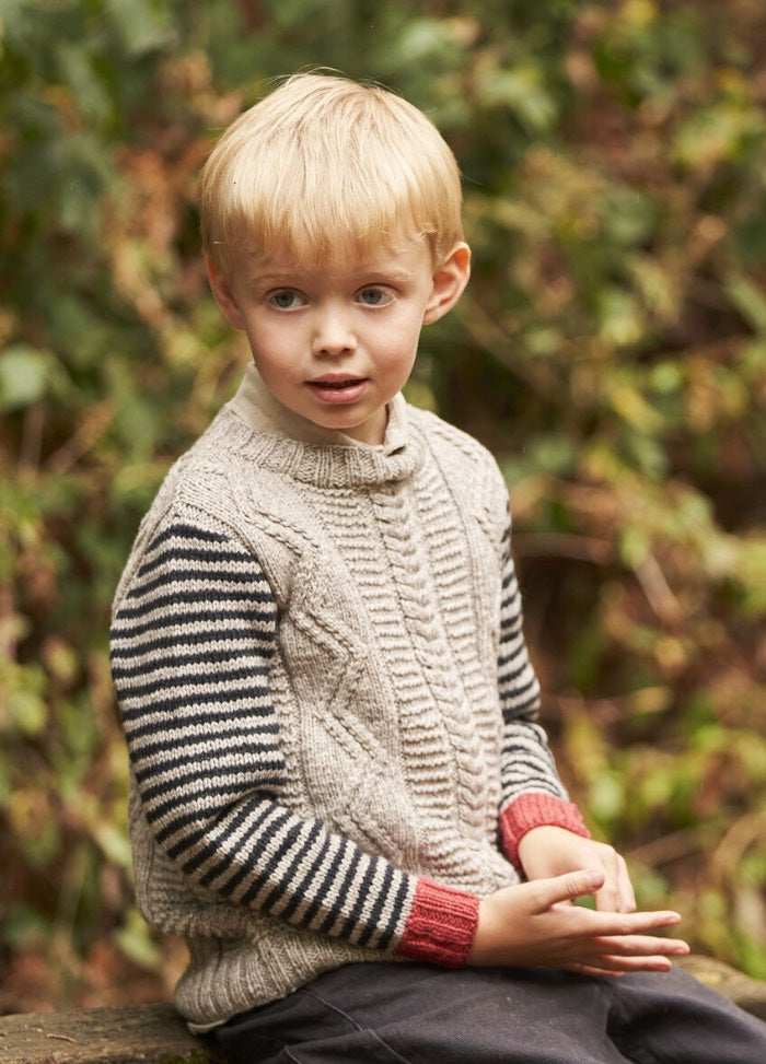 A young boy with blond hair sits outdoors. He wears a beige knit sweater with navy striped sleeves and red cuffs. The background is a blurred garden.