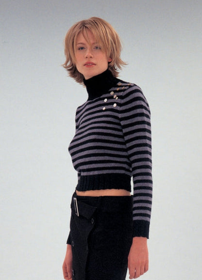 A person with short, layered blonde hair wears a black and gray striped turtleneck sweater with button details, paired with black pants. The expression is neutral.