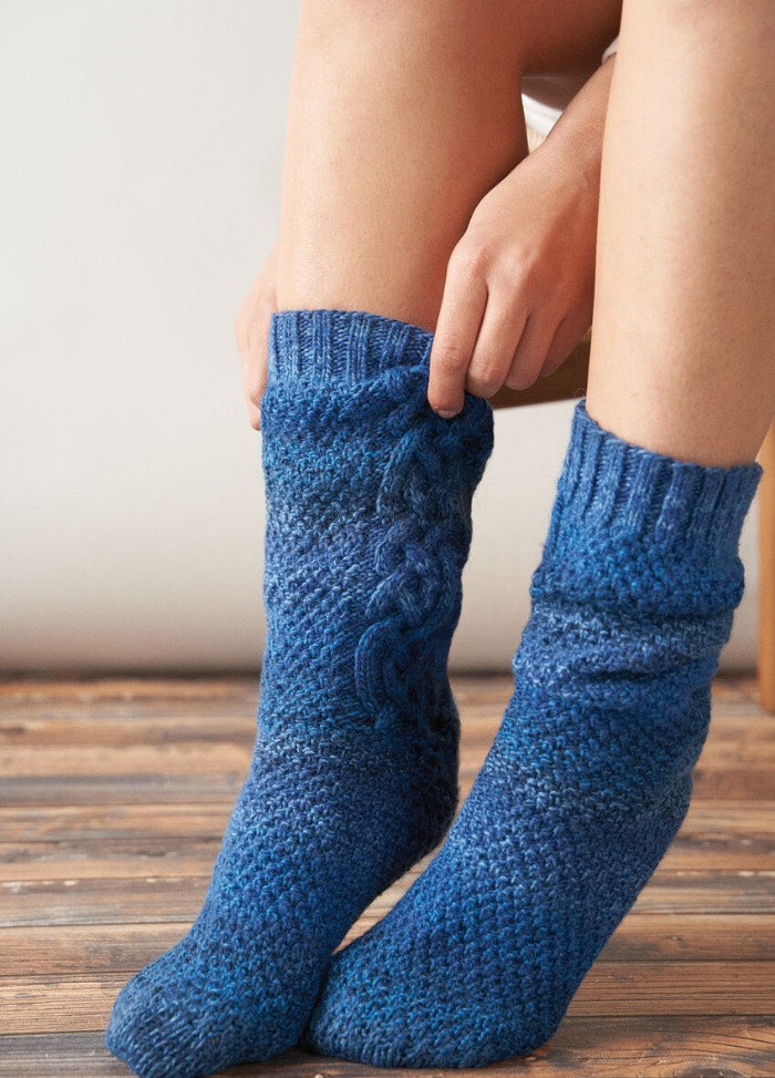 Close-up of a person wearing hand-knitted blue socks with intricate cable patterns, sitting on a wooden floor. The scene feels cozy and warm.