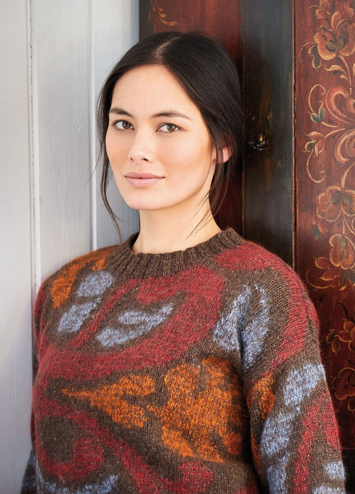 A woman with dark hair and an orange, brown, and blue patterned sweater leans against a wooden wall with ornate floral designs, offering a soft smile.